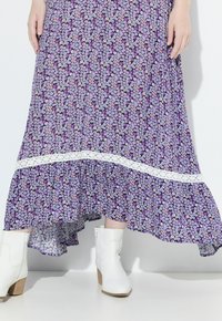 Abito maxi floreale viola con rifiniture in pizzo bianco, caratterizzato da un orlo svasato. Indossato con stivaletti bianchi alla caviglia e un tessuto testurizzato.