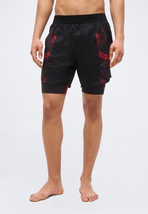 Férfi fekete sportnadrágot visel, piros kígyómintával és beépített kompressziós rövidnadrággal, mezítláb áll fehér háttéren.