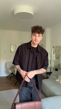 Borsa in pelle marrone con una lunga tracolla, caratterizzata da un design strutturato e cuciture in vista. La persona indossa una maglietta nera e jeans.