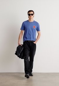 Blue cotton t-shirt with a red circular logo, paired with black jeans and a black leather jacket, worn with black sunglasses and shoes.