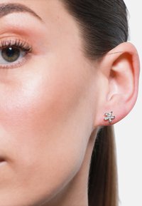 Sølv blomstformet stud earring med fem kronblader, prydet med små klare edelsteiner, satt mot en lys hudtone og jevn bakgrunn.