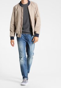 Světlobéžová bomber bunda s tmavě modrými žebrovanými manžetami, šedý svetr, vybledlé modré džíny a šedé tenisky. Model stojí vestoje.