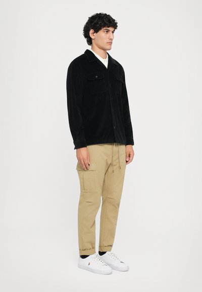 Black corduroy shirt jacket with two chest pockets, paired with khaki cargo pants. White sneakers complete the outfit.