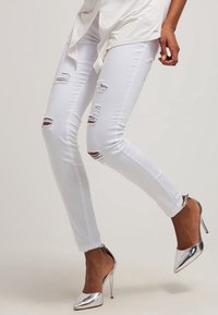 Jeans de mezclilla blanca ajustados con detalles desgastados, que presentan múltiples roturas. Combinados con zapatos de tacón alto plateados y brillantes.