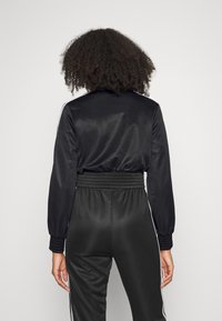 Kvinna med lockigt hår iförd en svart långärmad träningsjacka och matchande högmidjade byxor med vita sidostripes, vänd bort.