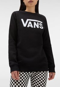 Nainen, joka on pukeutunut mustaan Vans-huppariin, jossa on valkoinen logo, ja mustavalkoisiin ruutuhousuihin, seisoo yksinkertaista taustaa vasten.