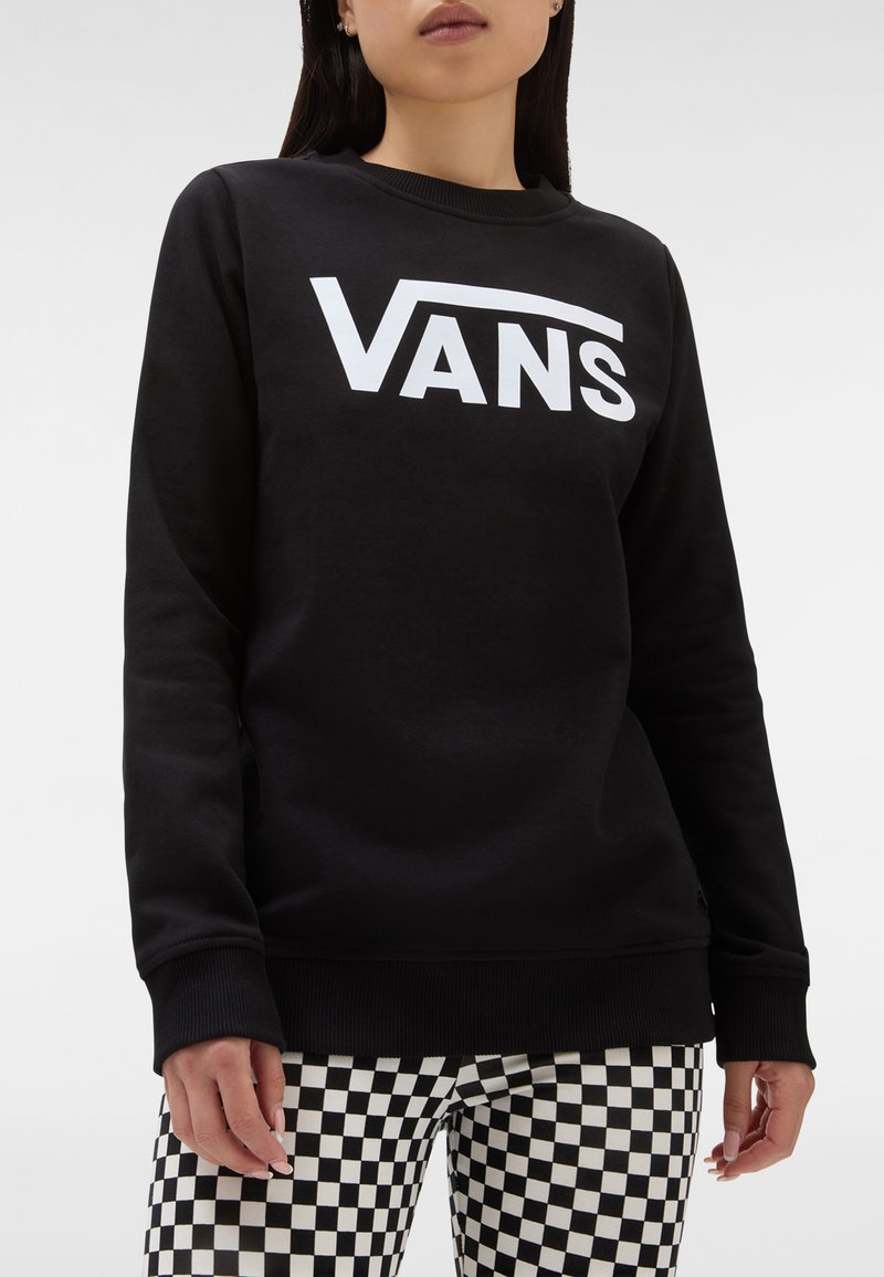 Nainen, joka on pukeutunut mustaan Vans-huppariin, jossa on valkoinen logo, ja mustavalkoisiin ruutuhousuihin, seisoo yksinkertaista taustaa vasten.