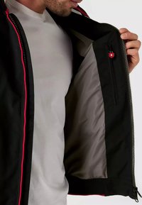 Veste noire avec un accent rouge, fermeture éclair à l'avant et poche intérieure. Doublée de tissu gris, elle présente une texture lisse.