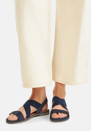 Tmavomodré sandále na plochom podpätku s remienkami, s prekríženým dizajnom a štruktúrovaným vrchom, nosené s krémovými širokými nohavicami.