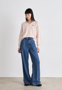 Pull en V rose clair, jean large bleu ample, et chaussures noires. Le pull présente le logo Lacoste.