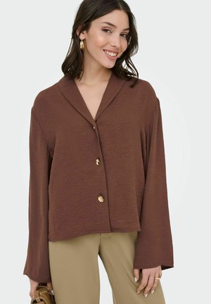 Jauna moteris šypsosi, dėvi laisvą rudą su sagomis palaidinę, smėlio spalvos kelnes, auksinius lankelio formos auskarus ir laiko mažą rudą rankinę.