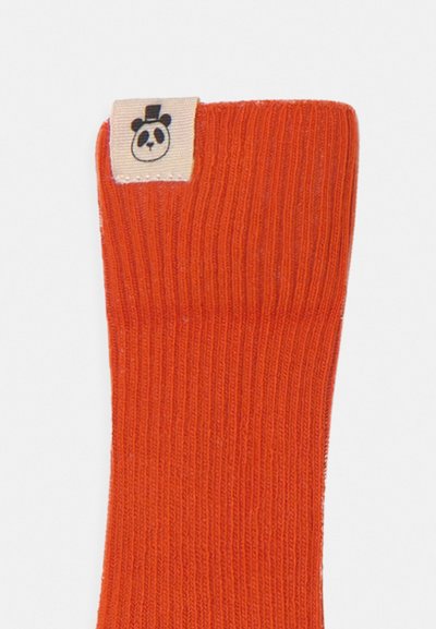Meia laranja canelada com uma etiqueta têxtil que apresenta um desenho de panda com um chapéu. O tecido parece ser macio e elástico.