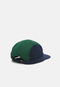 Gorra bicolor con una sección superior verde y un visor y parte trasera azul oscuro. Fabricada con un tejido texturizado y un panel de malla para mayor transpirabilidad.