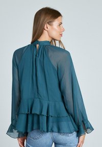 Blusa in chiffon color teal con maniche lunghe, orlo arricciato e dettaglio a taglio a chiave sulla schiena. Texture morbida con design a strati. Indossata con jeans blu.