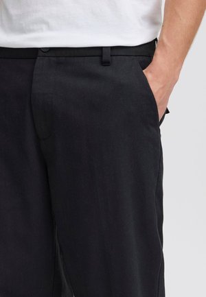 Person trägt schwarze Hose, eine Hand in der rechten Tasche, dazu ein weißes Hemd, das an der Taille sichtbar ist.