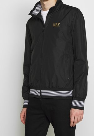 Homme portant une veste noire zippée avec des poignets et un ourlet rayés, logo doré EA7 Emporio Armani sur la poitrine et une chemise blanche en dessous.