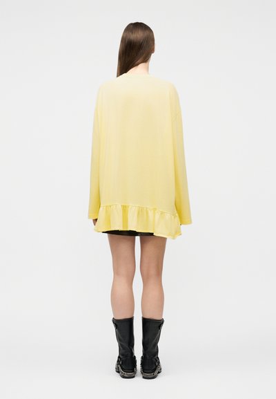 Γυναίκα που στέκεται με την πλάτη προς τα εμπρός, φοράει έναν χαλαρό κίτρινο μακρυμάνικο τοπ με βολάν στο τελείωμα, μαύρα σορτς και μαύρα μποτάκια μέχρι τη μέση της γάμπας, σε λευκό φόντο.