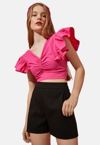 Rosa Cropped-Top mit gerafftem Vorderteil und Rüschensleeves, kombiniert mit hochTaillierten schwarzen Shorts. Glatte Stoffstruktur.