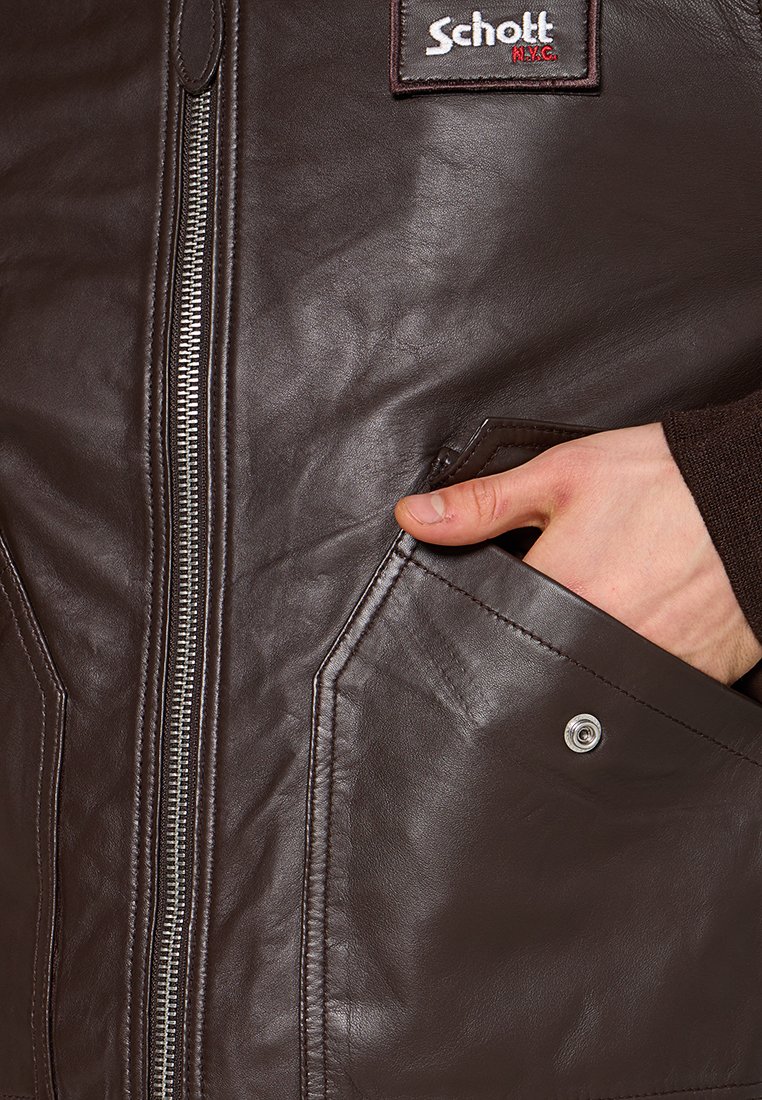 Veste en cuir marron avec fermeture éclair argentée et poche à bouton-pression, main partiellement dans la poche, logo Schott NYC visible.