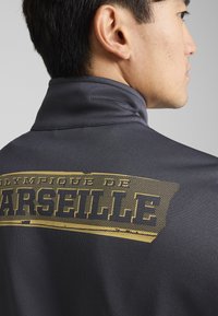 Grå zip-up jacka med hög krage, prydd med en stor guldig "Olympique de Marseille" grafi på baksidan. Slät struktur och figurnära design.