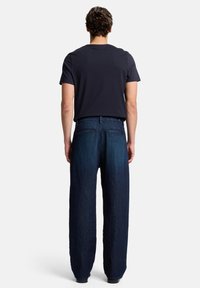 Jeans in denim blu scuro con vestibilità rilassata e gambe larghe; caratterizzati da due tasche posteriori e una semplice cintura; abbinati a una t-shirt nera.