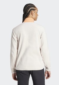 Chaqueta de forro polar beige claro con un cuello alto y hombros en un tono naranja claro que contrastan. Presenta mangas largas y una textura suave.