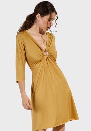 Frau mit lockigem Haar, die ein senfgelbes Kleid mit dreiviertel Ärmeln und einem runden Ausschnitt auf der Brust trägt und nach unten schaut.