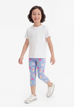 Παιδί που χαμογελάει και περπατάει, φοράει λευκό κοντομάνικο μπλουζάκι, μπλε κολάν με σχέδια παγωτού και λευκά αθλητικά παπούτσια.