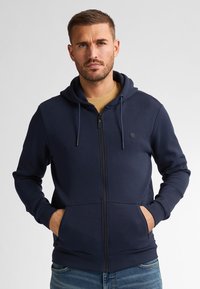 Navy zip-up hoodie made of soft fabric, featuring a drawstring hood, front pockets, and a small logo on the chest.