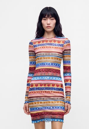 Femme aux longs cheveux noirs portant une robe bodycon rayée colorée avec du texte, des cœurs, des symboles de paix et des motifs en X sur fond blanc.