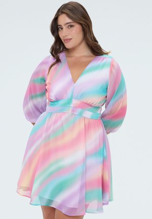 Femme aux longs cheveux bruns portant une robe courte à rayures pastel arc-en-ciel, manches bouffantes, col en V, taille ceinturée, et se tient avec confiance.