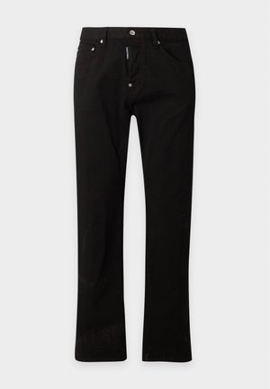 Giovane uomo con canotta nera, pantaloni neri con cintura e scarpe nere che sta davanti a uno sfondo bianco semplice.