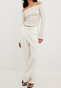 Kvinna i beige ribbad långärmad off-shoulder topp, högmidjade lösa vita byxor och svarta öppna klackar med vit nagellack.