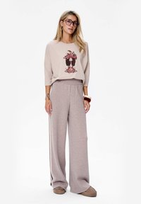 Vrouw in een beige langwerpig shirt met grafische print, taupe wijduitlopende broek met zijstrepen, beige instappers, grote brillen en creolen.