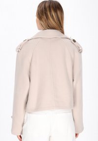 Lys beige jakke med en struktureret krave, militærstil skulderepauletter og en kortet bund. Fremstillet af glat, tekstureret stof.