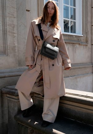 Kvinne i beige trenchcoat og vide bukser poserer på en steinkant utenfor en bygning, bærende en svart crossbody-veske med bred stropp.