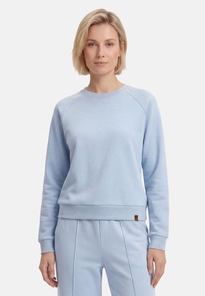 Blonde vrouw met lichtblauwe trui en bijpassende broek, staande tegen een effen witte achtergrond, kijkt recht vooruit met een neutrale gezichtsuitdrukking.