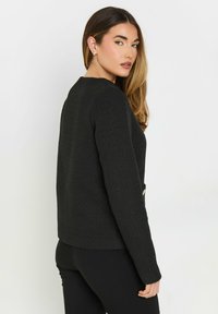 Musta teksturoidumpaita, jossa on pitkät hihat ja muodollinen istuvuus. Ominaisuuksiin kuuluu pyöreä kaula-aukko ja kaksi koristeellista etutaskua.