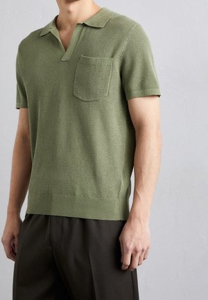 Mann trägt ein kurzärmeliges, strukturiertes olivgrünes Strickpolohemd mit Brusttasche und dunkle Hose vor einfachem hellem Hintergrund.