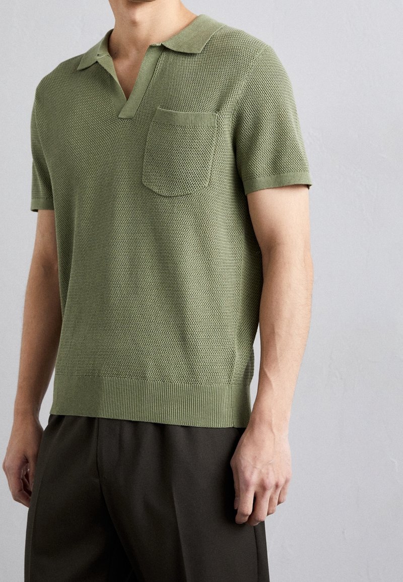 Homme portant un polo en maille texturée vert olive à manches courtes avec une poche poitrine et un pantalon foncé, sur un fond clair uni.