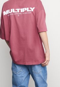 Camiseta oversized de color burdeos hecha de algodón, con gran texto blanco "MULTIPLY APPAREL CLUB 2024" en la parte posterior. Diseño casual, mangas cortas.