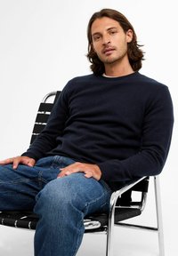Navyblauwe trui met ronde hals en lange mouwen, gecombineerd met lichtblauwe denim jeans. Zit op een zwart en zilver gekleurde stoel.