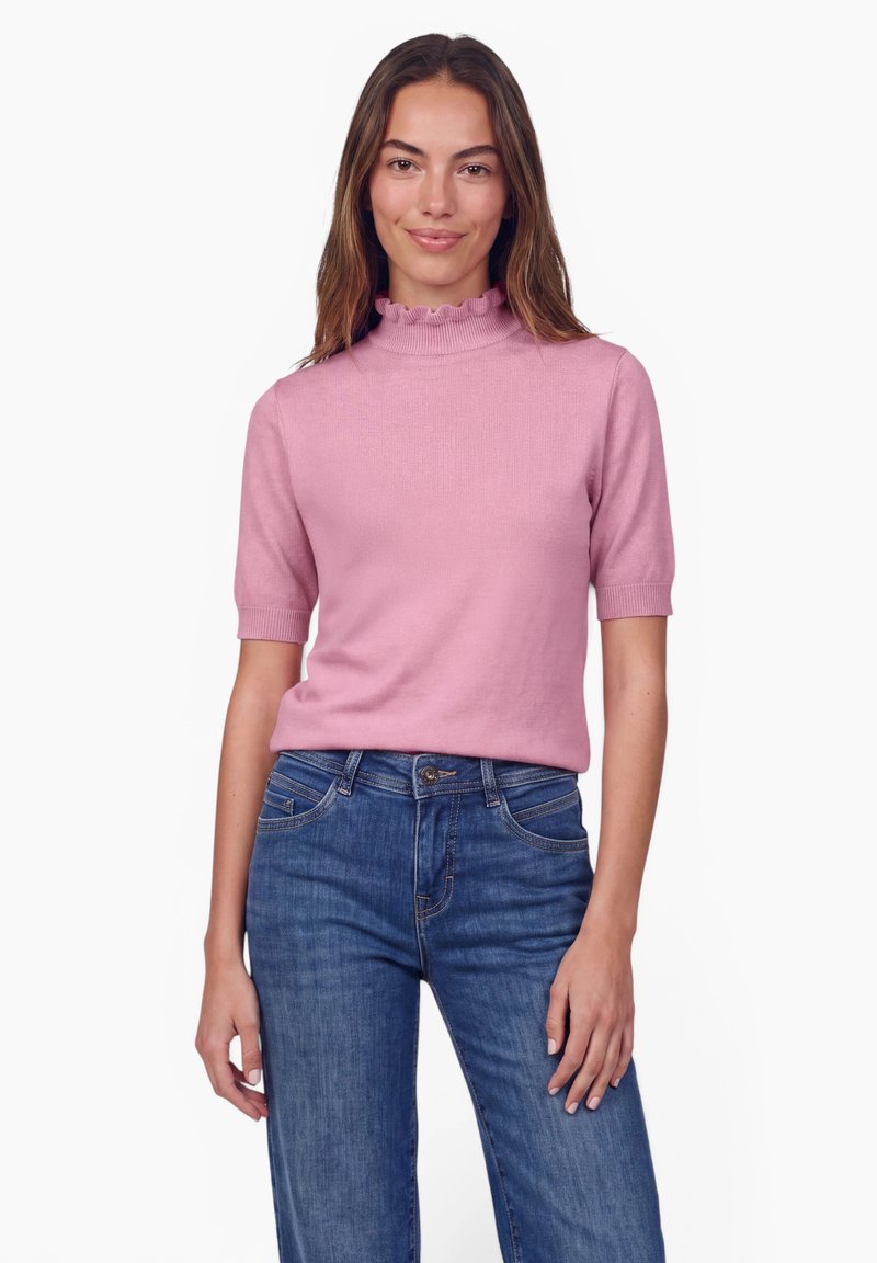 Femme portant un haut rose clair à manches courtes avec un col haut à volants et un jean bleu, debout devant un fond blanc uni.