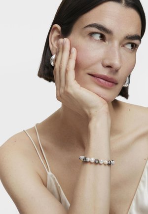 Pulsera de perlas y piedras preciosas con acentos de plata, que presenta cuentas redondas de diferentes tamaños y texturas. Llevada en una muñeca, con un fondo de tela suave.
