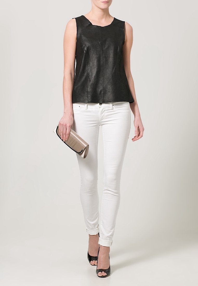 Femme portant un haut noir sans manches, un jean skinny blanc, des talons ouverts noirs, et tenant une pochette beige.