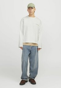Sweatshirt lung gri cu mâneci lungi, suprapus peste un top bej, asortat cu blugi ușor largi de culoare albastră și cizme maro cu șireturi. O cușma verde completează ținuta.