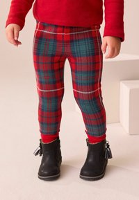 Niña vestida con leggings de cuadros rojos y verdes, calcetas rojas, botas negras hasta el tobillo con flecos, y una camiseta roja de manga larga, de pie sobre un suelo claro.