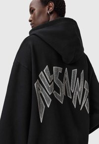 Zwarte oversized hoodie met een zilveren rhinestone-logo op de achterkant, gemaakt van een gladde, gestructureerde stof.