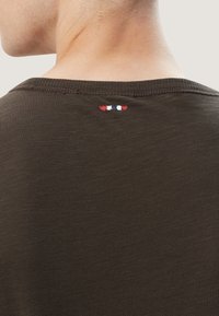 Dunkelbraunes T-Shirt mit Rundhalsausschnitt, das ein kleines, gesticktes Detail in Rot, Weiß und Blau am hinteren Kragen aufweist. Weicher, strukturierten Stoff.