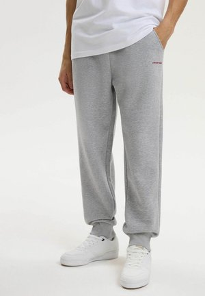 Osoba ubrana w jasnoszare dresowe spodnie i białe trampki, stojąca z jedną ręką w kieszeni na jednolitym tle.